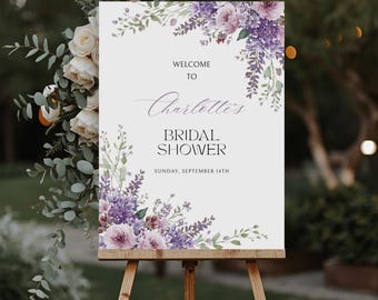 May include: A white sign on a wooden easel, decorated with purple and lavender floral arrangements. The sign reads "Welcome to Charlotte's Bridal Shower" with the date "Sunday, September 14th".