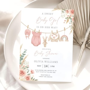 May include: A baby shower invitation card featuring a clothesline with a pink bonnet, pink romper, plush rabbit, bib, and rainbow toy. Floral accents in pink and green frame the text A SWEET Baby Girl IS ON HER WAY! and event details.