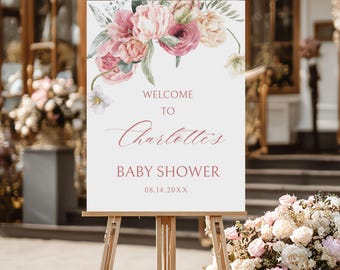 May include: A welcome sign for a baby shower, displayed on a wooden easel. The sign features floral arrangements in pink, peach, and white. Text reads "Welcome to Charlotte's Baby Shower" with a date below.
