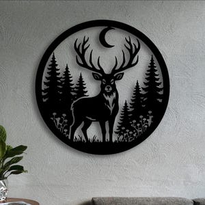 May include: A circular black metal wall art piece featuring a detailed silhouette of a stag with large antlers standing in a forest of pine trees. Above the stag, a crescent moon is visible, and small floral elements are at the base of the scene.