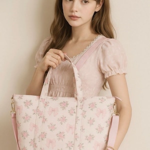 Puede incluir: Un bolso tote rosa claro con un estampado floral y de lazos. El bolso tiene una correa para el hombro y asas rosas a juego. Los herrajes dorados acent&uacute;an el bolso. El bolso lo sostiene una persona que lleva un vestido y una diadema rosas.