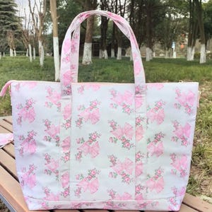 Puede incluir: Un bolso tote grande con un fondo gris claro y un patr&oacute;n repetido de lazos rosas y detalles florales. El bolso tiene asas rosas a juego y un cierre de cremallera. El bolso est&aacute; sobre una superficie de madera.