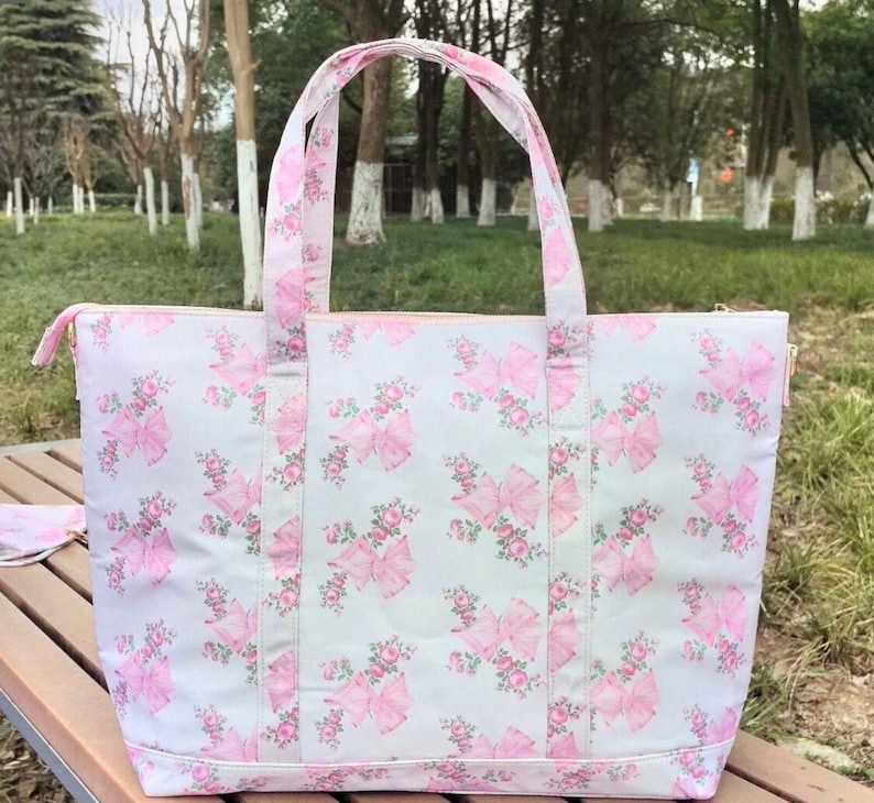 Puede incluir: Un bolso tote grande con un fondo gris claro y un patr&oacute;n repetido de lazos rosas y detalles florales. El bolso tiene asas rosas a juego y un cierre de cremallera. El bolso est&aacute; sobre una superficie de madera.