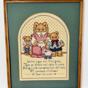 May include: Cross-stitch artwork in a wooden frame. The image features a family of teddy bears in a cross-stitch design. The bears are dressed in various outfits. Text at the bottom reads: "Little boys and little girls, Toys on wheels and dolls in curls..."