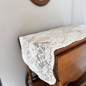May include: A vintage wooden tea cart with a cream-colored lace tablecloth. The cart has a warm, brown finish and features a drop-leaf design. The lace tablecloth has a floral pattern.