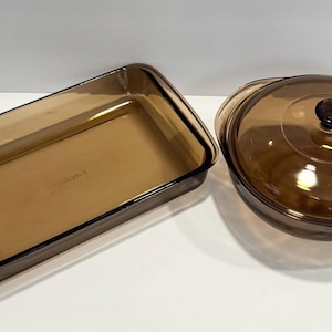 May include: Two amber-colored glass cookware pieces. One is a rectangular baking dish, and the other is a round casserole dish with a lid. Both pieces are made of transparent glass, allowing visibility of the contents. Ideal for baking and serving.