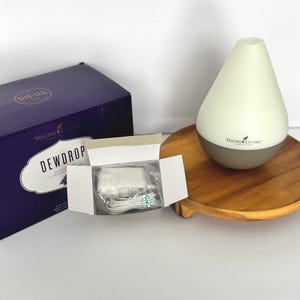 May include: A white and gray essential oil diffuser with the text "Young Living" on a wooden tray. A purple box and a white power adapter are also visible. The box reads "DEWDROP" and "Young Living".