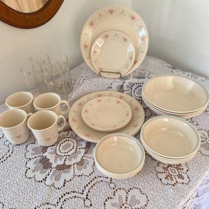 May include: A cream-colored dinnerware set with pink heart accents. The set includes plates, bowls, cups, and glasses. The plates and bowls have a pink and green trim. The glasses and cups have heart designs.