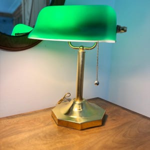 May include: A green-shaded desk lamp with a brass base and stem. The lamp has a pull chain and sits on a wooden surface. The shade is a vibrant green, and the base is a gold color.