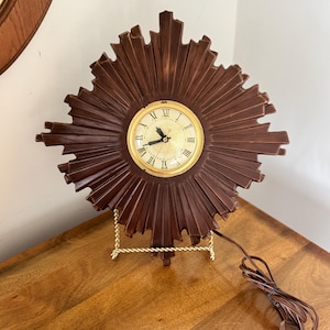 May include: A vintage sunburst clock with a dark brown wooden frame and a gold-colored clock face. The clock has Roman numerals and is displayed on a gold-colored stand. A brown electrical cord is attached.