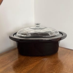 May include: A black ceramic oval-shaped slow cooker insert with a wide rim and a clear glass lid featuring a central knob handle. The vessel has a smooth, glossy finish and a deep basin, designed for use within a heating base unit.