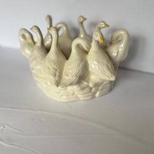 May include: Cream-colored ceramic centerpiece featuring a ring of stylized swans with yellow beaks. The swans encircle a central bowl, creating a decorative and elegant design. The base has a woven basket texture.