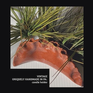 May include: A vintage, handmade candle holder in a rust-colored finish. The holder has a unique, organic shape and is accented with green and yellow faux pine needles. Text on the image reads "VINTAGE, UNIQUELY HANDMADE IN PA., candle holder."