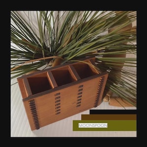 Puede incluir: Un organizador de madera con cuatro compartimentos, con acabado de madera natural y detalles en marrón oscuro. El diseño incluye ranuras verticales en los laterales y un patrón decorativo. La palabra "MOONSPON" es visible.