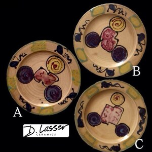 May include: Three handmade ceramic plates with abstract designs. Each plate features a beige background with colorful, geometric shapes in shades of purple, yellow, and red. The rims are decorated with a wavy pattern and the text "D. Lasser Ceramics" is visible.