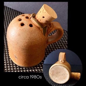 May include: A brown ceramic bottle with a cork stopper. The bottle has a handle and several holes on top. The text "circa 1980s" is visible. The bottle is likely a decorative item or a unique container.
