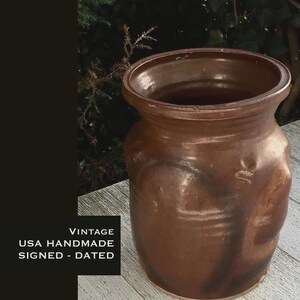 May include: A vintage, handmade brown ceramic vase with a wide opening and a textured surface. The vase has a rounded body and a slightly flared rim. The text "VINTAGE USA HANDMADE SIGNED - DATED" is visible.
