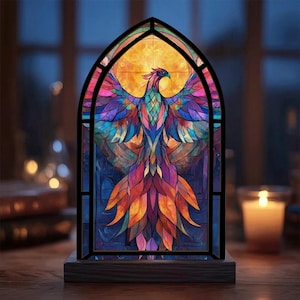 May include: A stained glass window depicting a colorful phoenix with outstretched wings. The bird is set against a yellow background, framed by a black border. The window is mounted on a dark wooden base, creating a decorative accent piece.