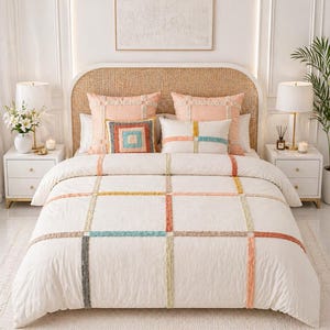 Puede incluir: Una cama con cabecero tejido, adornada con almohadas color melocotón y un edredón blanco. El edredón presenta un patrón de cuadrícula con acentos coloridos y texturizados en naranja, azul, amarillo y gris. Dos mesitas de noche blancas flanquean la cama, cada una con una lámpara.