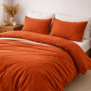 Puede incluir: Una cama con una funda nórdica naranja y fundas de almohada a juego. La ropa de cama está colocada sobre un colchón blanco, con una mesita de noche de madera y una pequeña planta en el fondo. La habitación tiene una paleta de colores neutros.