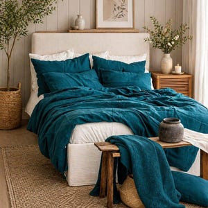 May include: A teal bedspread and pillow set on a white bed frame. The bedroom features a woven rug, a wooden bench, and a framed botanical print. A wicker basket and a vase with flowers add to the room's decor.