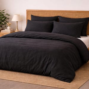 Puede incluir: Un juego de cama negro con funda nórdica y fundas de almohada sobre una cama con cabecero tejido. La cama está sobre una alfombra beige, con mesitas de noche y lámparas a ambos lados.