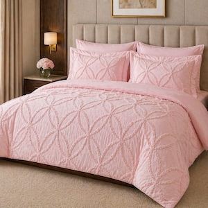 Pode incluir: Um conjunto de colcha e almofadas rosa claro. A roupa de cama apresenta um design de padrão circular em relevo. A cama é composta por duas fronhas e um edredom. A cama está contra uma cabeceira e uma parede de tons neutros.