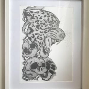 May include: Framed black and white art print. The illustration depicts a jaguar's head with an open mouth, teeth bared, above two skulls. The artwork is presented in a white frame.