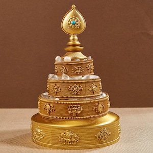 May include: A gold-coloured, tiered decorative object with detailed embossed patterns and a turquoise accent. The object has a teardrop-shaped top and is adorned with clear crystals. The base is round and the overall design is ornate.