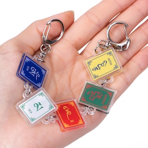 May include: A multi-colored keychain featuring five rectangular acrylic charms in blue, white, red, green, and yellow. Each charm displays a unique symbol and text, including OM, MA, and HUM, connected by silver metal jump rings and a lobster clasp.