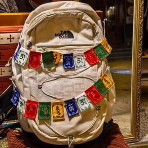 Peut inclure: Un sac à dos beige avec le logo The North Face, orné d'une guirlande de drapeaux de prière colorés. Les drapeaux rectangulaires présentent diverses couleurs et symboles. Le sac à dos est exposé dans une boutique avec un miroir reflétant d'autres articles.