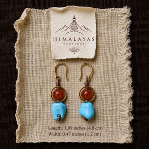 May include: A pair of drop earrings featuring a gold-tone metal ring, a round reddish-brown bead, and a light blue turquoise-coloured stone. The earrings have a length of 4.8 cm and a width of 1.2 cm, with text reading HIMALAYAS BOUTIQUE.