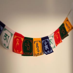 K&ouml;nnte beinhalten: Eine Reihe rechteckiger Gebetsfahnen in leuchtenden Farben wie Rot, Gr&uuml;n, Gelb, Blau und Wei&szlig;. Jede Fahne ist mit tibetischer Schrift und Symbolen bedruckt, darunter die Texte PADME, HUM und OM, die in einer horizontalen Anordnung h&auml;ngen.