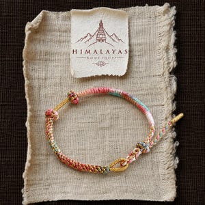 May include: A braided cord bracelet featuring a mix of pink, blue, and gold threads with decorative knots and a gold-toned loop closure. Above the bracelet is a rectangular paper tag with the text Himalayas Boutique and a line art illustration.