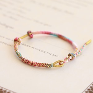 May include: A colorful woven bracelet with pink, blue, and gold threads. The bracelet features a braided design with gold-colored metal accents at the clasp and ends. The bracelet is on a light-colored background.