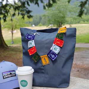 K&ouml;nnte beinhalten: Eine schieferblaue Tragetasche, verziert mit einer Kette aus bunten Gebetsfahnen, eine hellblaue Baseballkappe mit Patagonia-Logoaufn&auml;her und ein wei&szlig;er Einweg-Kaffeebecher mit einem gr&uuml;nen Whole Foods Market-Logo auf der Vorderseite.