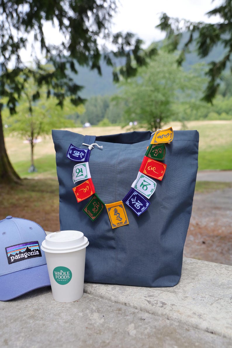 K&ouml;nnte beinhalten: Eine schieferblaue Tragetasche, verziert mit einer Kette aus bunten Gebetsfahnen, eine hellblaue Baseballkappe mit Patagonia-Logoaufn&auml;her und ein wei&szlig;er Einweg-Kaffeebecher mit einem gr&uuml;nen Whole Foods Market-Logo auf der Vorderseite.