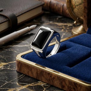May include: A silver ring featuring a large, rectangular, polished black stone set in the center. The band has a smooth, metallic finish with small, clear, sparkling stones embedded along the sides of the setting, creating a refined appearance.