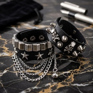 May include: Two black leather wristbands featuring silver-tone metal hardware. One band has rectangular studs, star-shaped rivets, and dangling chains. The other band features skull and crossbones emblems, round studs, and conical spikes.
