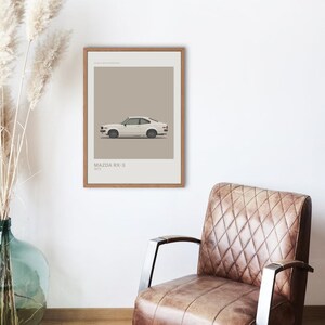 Könnte beinhalten: Ein gerahmter Druck mit einem weißen Mazda RX-3 von 1972. Das Auto ist in einem minimalistischen Stil vor einem neutralen Hintergrund dargestellt. Der Druck befindet sich in einem Holzrahmen und wird an einer weißen Wand in der Nähe eines braunen Ledersessels ausgestellt.