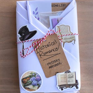 May include: A white package tied with red and white twine, adorned with stickers of a top hat, armchair, and book. A brown tag reads "Historical Romance - Mystery Abounds."