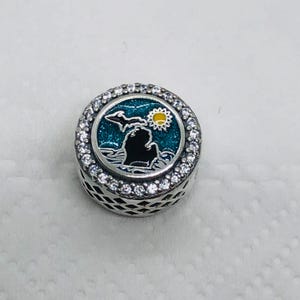 Peut inclure: Un charm en argent avec un design rond représentant une silhouette noire d'un état, un soleil jaune et un fond turquoise. Le charm est incrusté de petites pierres claires sur le bord.