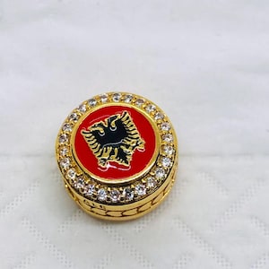 May include: A gold-colored ring with a red center featuring a black double-headed eagle emblem. The ring is encrusted with small, clear gemstones around the red emblem. The ring has a detailed, textured band.