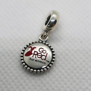 Puede incluir: Un charm de metal plateado con un colgante circular de borde perlado. El centro muestra un icono de vestido en esmalte rojo y el texto negro Go Red for women TM. El charm incluye una anilla de plata pulida para sujetarlo a una pulsera o cadena.