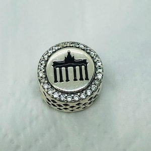 Könnte beinhalten: Ein silberner Charm mit einer schwarzen Silhouette des Brandenburger Tors auf weißem Hintergrund. Der runde Charm hat einen Rand aus klaren, funkelnden Steinen. Die Seiten des Charms haben ein dekoratives Lochmuster.