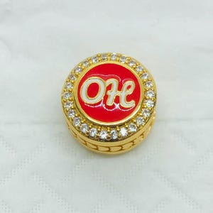 Peut inclure: Une bague dorée avec un centre rouge portant les lettres blanches "OH". La bague est entourée d'une rangée de pierres claires et scintillantes. La bague a une bande détaillée et texturée.