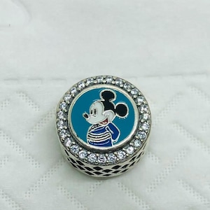 Op de afbeelding: Een ronde zilveren bedel met een centrale teal emaille inleg met een afbeelding van Mickey Mouse in een blauw-wit gestreept shirt. De bedel is omrand door een ring van heldere, sprankelende zirkonia steentjes en heeft een rasterpatroon.