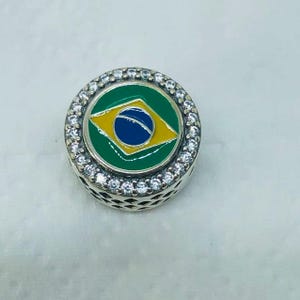 May include: A round charm featuring the Brazilian flag, with a green circle, yellow diamond, and blue globe design. The charm is surrounded by a ring of small, clear stones and has a silver-colored, openwork base.