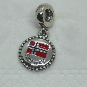 Könnte beinhalten: Ein silberner Charm mit einem Norwegen-Flaggen-Design. Der Charm hat einen runden Anhänger mit der norwegischen Flagge in Rot, Weiß und Blau und dem Wort "Norway" in Schwarz. Der Charm hat einen Perlenrand und eine Schlaufe zur Befestigung.