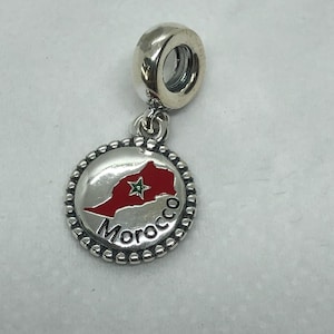 Peut inclure: Charm en argent avec un pendentif rond représentant une carte du Maroc rouge et verte. Le mot "Morocco" est écrit en noir. Le charm a une bordure perlée et une boucle argentée pour l'attache.